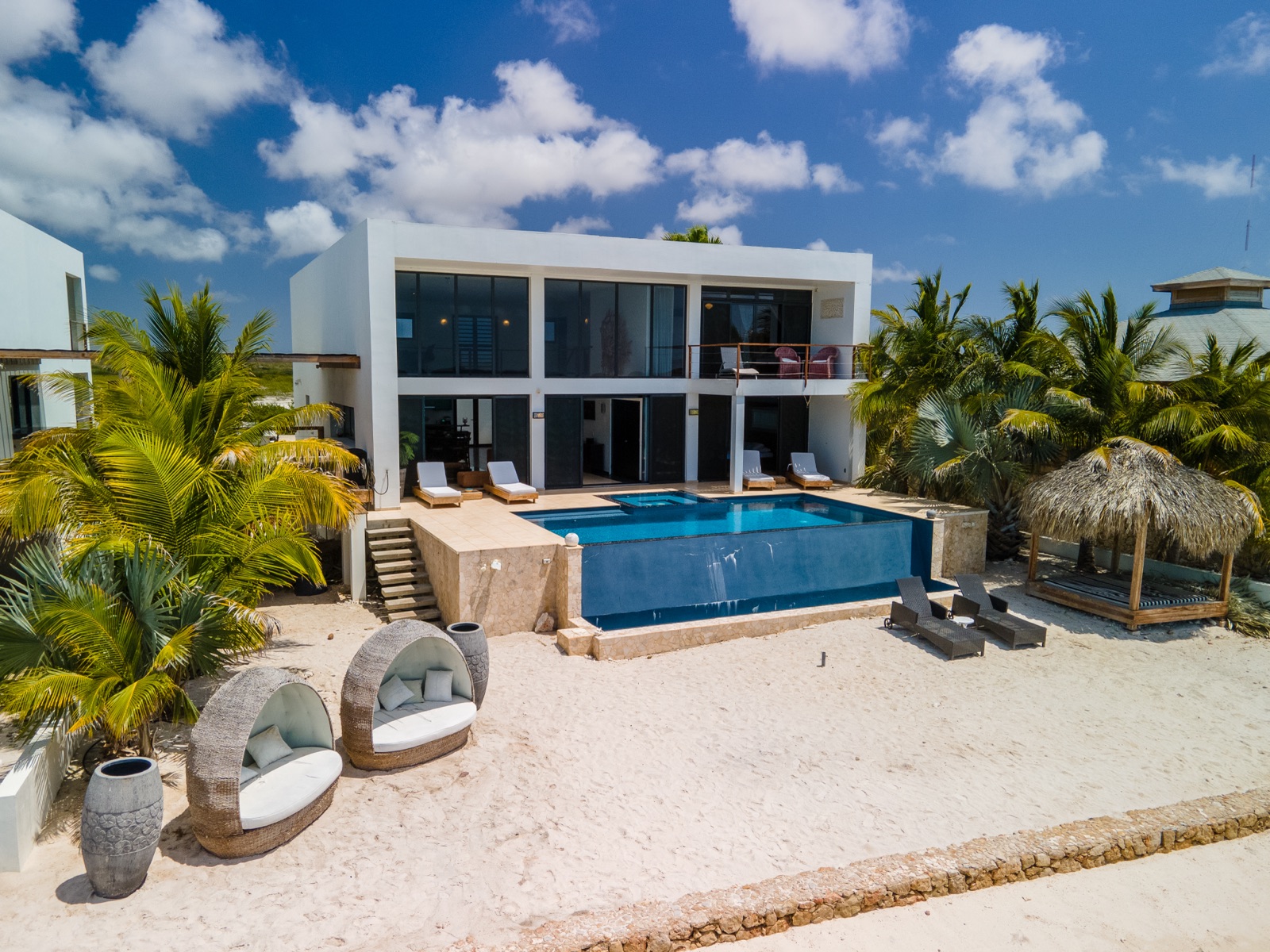 Luxury Bonaire villa with modern white architecture, a private infinity pool overlooking a sandy beach with palm trees and lounge cabanas.